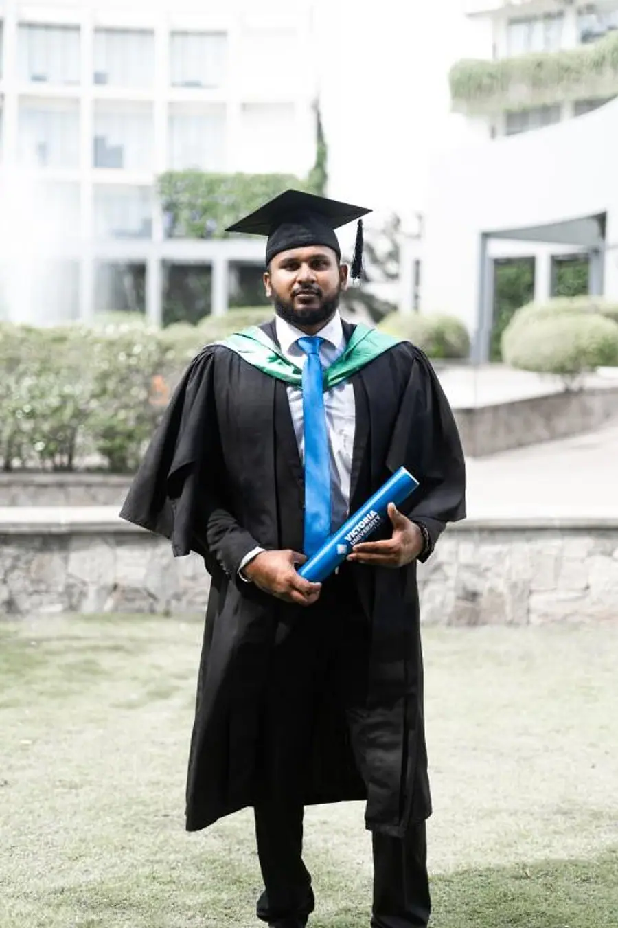 Dave Bowatta on graduation day in gown and mortarboard, holding a Victoria University degree tube