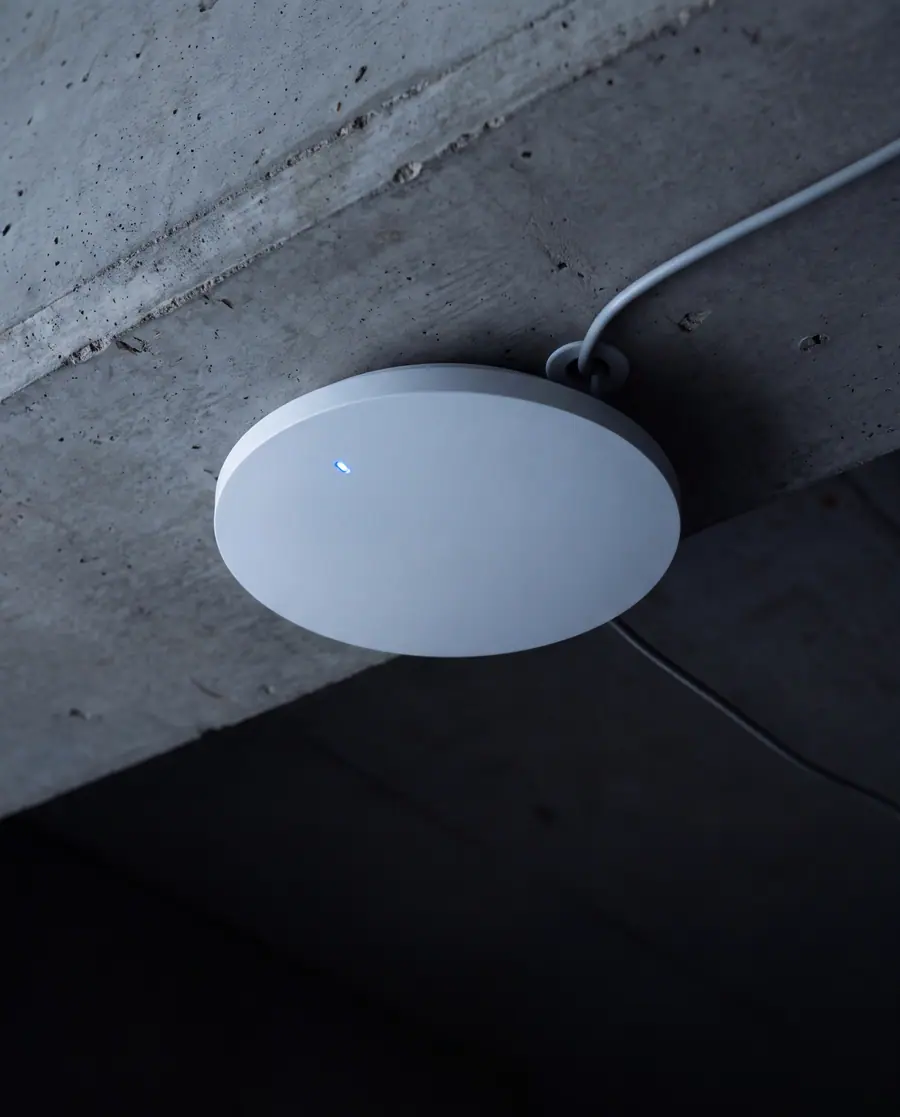 A white disc wireless access point on a board formed concrete soffit, seen from below, one cable dropping into it and a single blue status light.
