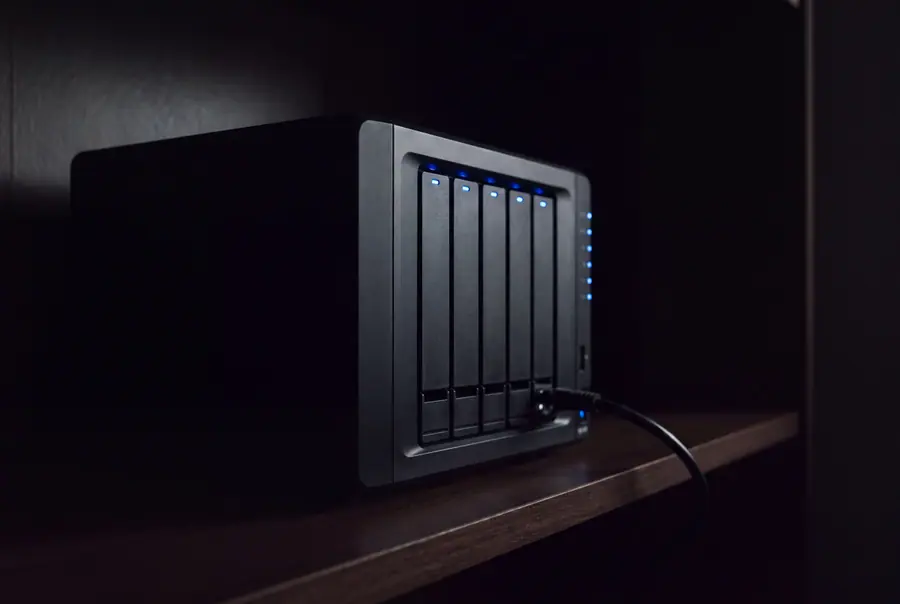 A five bay network attached storage unit on a shelf in a dark room, each bay lit blue, one network cable dressed into the back.