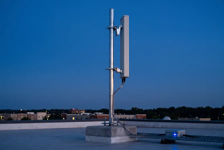 A panel antenna clamped to a galvanised pole on a concrete plinth on a flat roof at blue hour, its cable dressed down a tray into a bulkhead box.