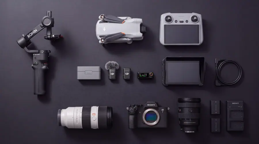 A gimbal, a folding drone and its controller, a wireless microphone kit, a field monitor, a camera body, lenses and batteries laid out on a dark surface.